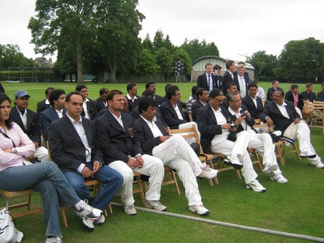 Players and guests in blazers seated on chairs at the edge of a ground